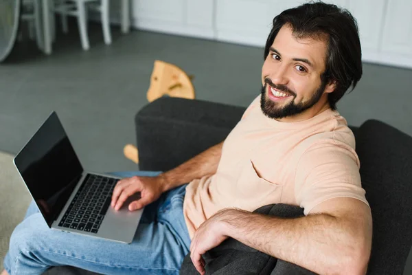 Vista lateral do homem sorridente com laptop descansando no sofá em casa — Fotografia de Stock