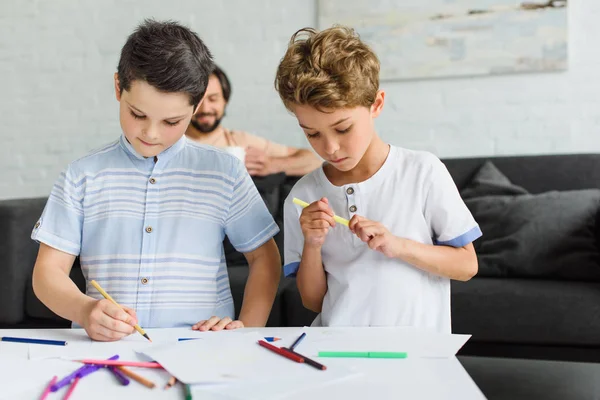 Niños dibujando dibujos con lápices de colores mientras padre descansa en el sofá en casa — Stock Photo