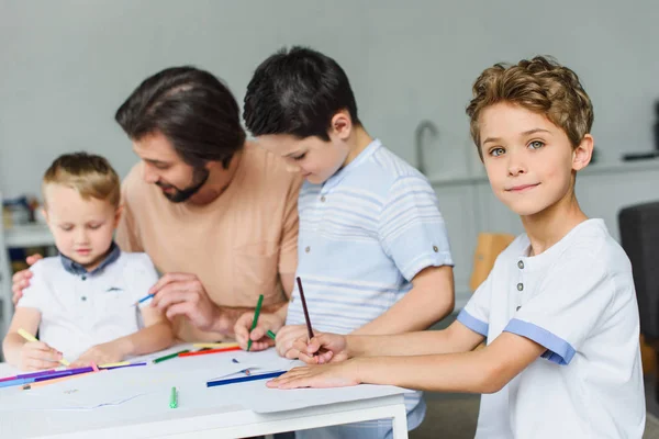 Селективное внимание отца помогает сыновьям рисовать картинки с цветными карандашами дома — стоковое фото