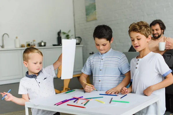 Messa a fuoco selettiva di bambini che disegnano immagini con matite colorate mentre il padre riposa sul divano di casa — Foto stock