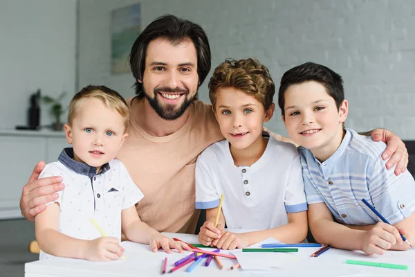 Портрет улыбающегося отца и сыновей за столом с бумагами и красочными карандашами дома — стоковое фото