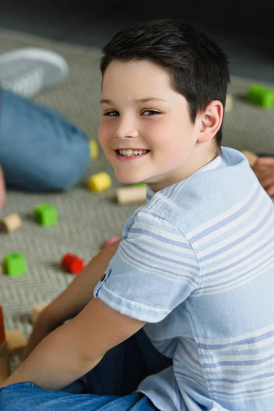 Side view of smiling boy looking at camera while sitting on floor with wooden blocks at home — Stock Photo