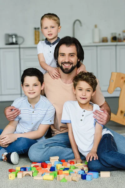 Happy father hugging little sons and looking at camera while sitting on floor with wooden blocks at home — Stock Photo