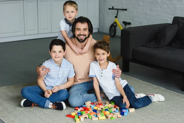Feliz padre abrazando a los hijos pequeños y mirando a la cámara mientras está sentado en el suelo con bloques de madera en casa - foto de stock