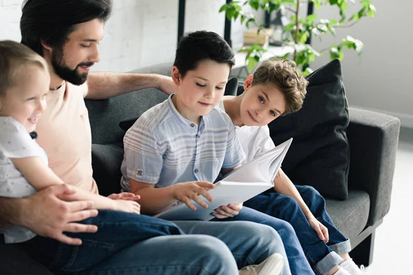 Vista lateral del padre y los hijos leyendo el libro juntos en casa - foto de stock