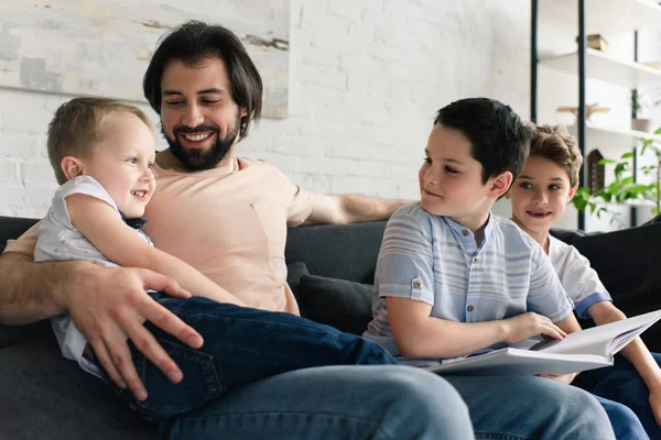 Happy father and sons resting on sofa while reading book together at home — Stock Photo