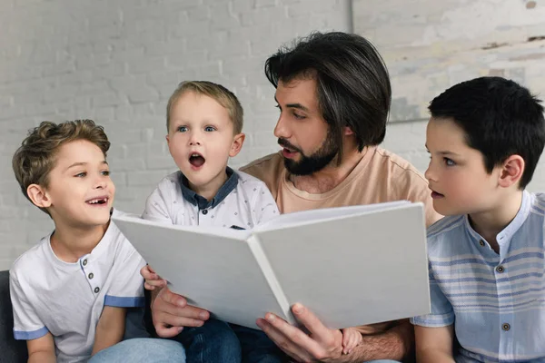 Retrato de padre e hijos pequeños leyendo el libro juntos en casa - foto de stock