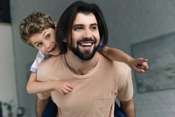 Retrato de pai feliz e filho pequeno apontando para longe enquanto piggybacking juntos em casa — Fotografia de Stock