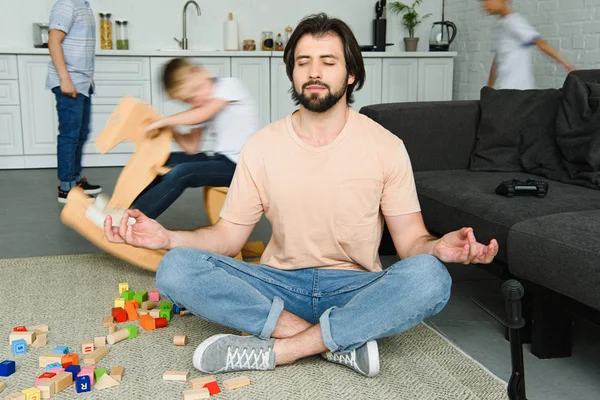 Напряженный отец сидит в позе лотоса на полу и сыновья играют вокруг в комнате — стоковое фото
