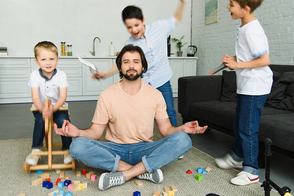 Pai estressado sentado em pose de lótus no chão e filhos brincando no quarto — Fotografia de Stock