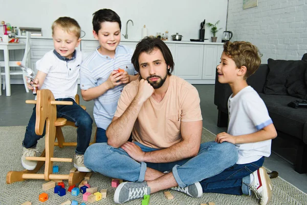 Padre estresado sentado en el suelo y los hijos jugando en la habitación - foto de stock