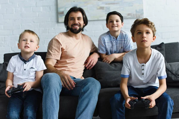 Sorrindo pai olhando para filhos pequenos sentados no sofá e jogando videogames juntos em casa — Fotografia de Stock