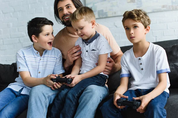 Lächelnder Vater mit Blick auf kleine Söhne, die zu Hause auf dem Sofa sitzen und gemeinsam Videospiele spielen — Stockfoto