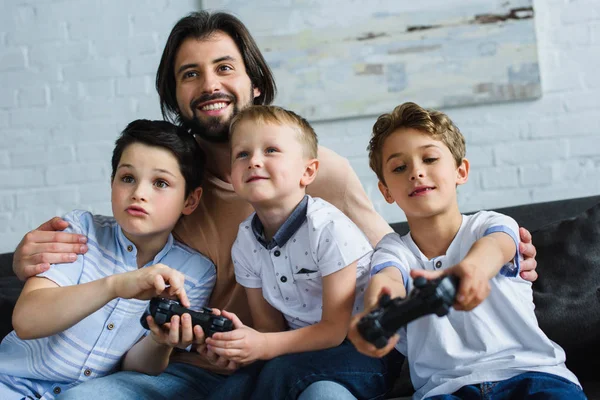 Padre sorridente guardando i piccoli figli seduti sul divano e giocare ai videogiochi insieme a casa — Foto stock