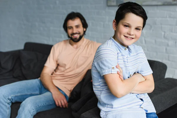 Enfoque selectivo del niño sonriente con los brazos cruzados y el padre descansando en el sofá detrás en casa - foto de stock