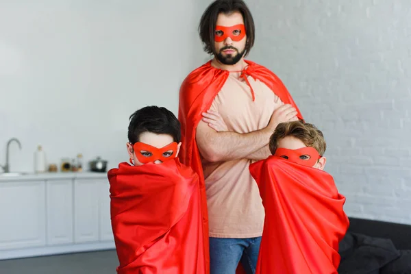 Retrato de homem e filhos em trajes de super-herói vermelho olhando para a câmera em casa — Fotografia de Stock