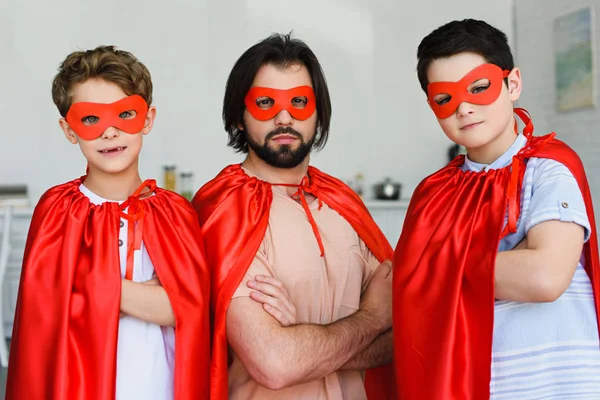 Retrato de padre e hijos en trajes de superhéroe rojo con los brazos cruzados mirando a la cámara en casa - foto de stock