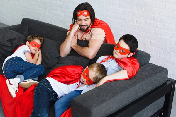 Visão de alto ângulo de pai feliz olhando para filhos adormecidos em trajes de super-herói vermelho no sofá em casa — Fotografia de Stock