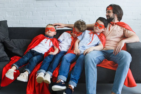 Père souriant assis près des fils endormis en costumes de super-héros rouge sur le canapé à la maison — Photo de stock