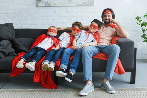 Smiling father sitting near sleeping sons in red superhero costumes on sofa at home — Stock Photo