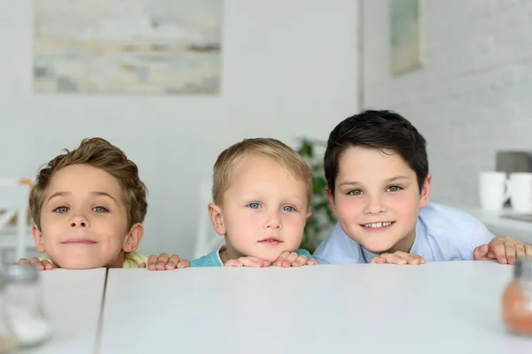 Niños pequeños escondidos detrás de la mesa y mirando a la cámara en casa - foto de stock