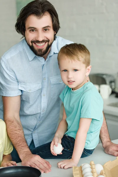 Portrait of smiling father and little son with raw egg in hand in kitchen at home — Stock Photo
