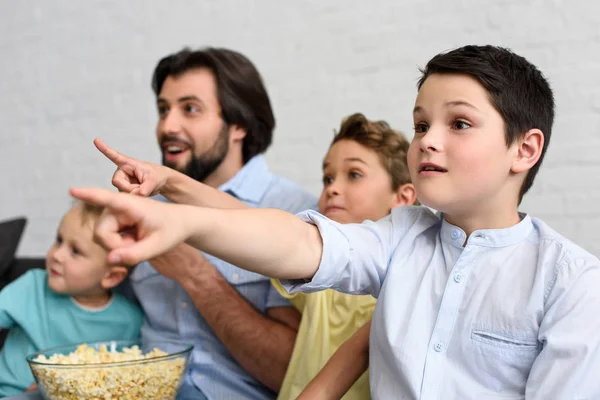 Возбужденный мужчина и маленькие сыновья с попкорном указывая на то, во время просмотра фильма вместе дома — стоковое фото
