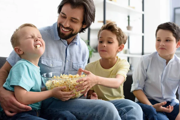 Улыбающийся мужчина и маленькие сыновья едят попкорн, смотря фильм вместе дома — стоковое фото