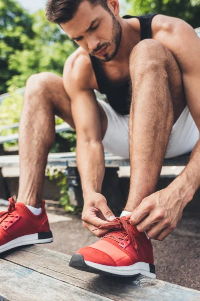 Serio giovane sportivo allacciatura lacci delle scarpe mentre seduto sulla panchina al parco giochi sportivo — Foto stock