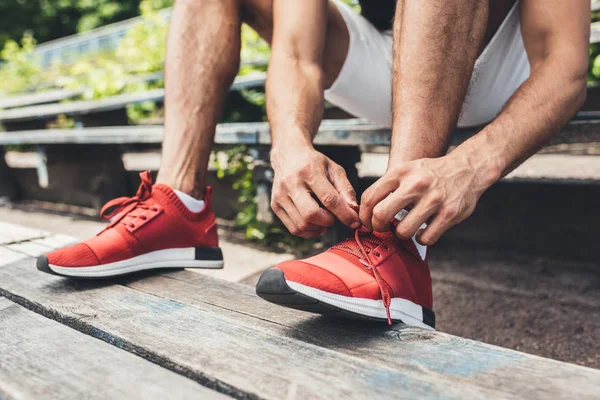 Immagine ritagliata di sportivo allacciatura lacci delle scarpe mentre seduto sulla panchina al parco giochi sportivo — Foto stock