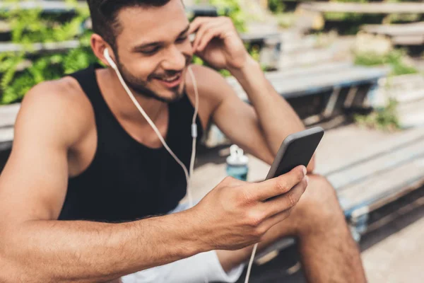 Sonriente joven escuchando música con smartphone y auriculares en el banco - foto de stock