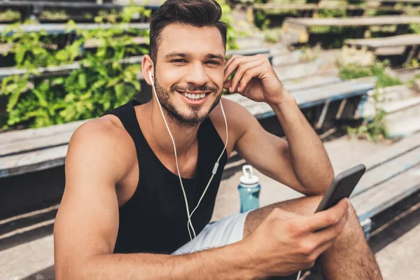 Красивый молодой человек слушает музыку со смартфоном и наушниками на скамейке — стоковое фото