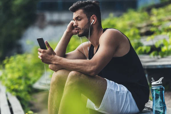 Deportista enfocado con botella de agua escuchando música con smartphone y auriculares en el banco en el parque deportivo - foto de stock