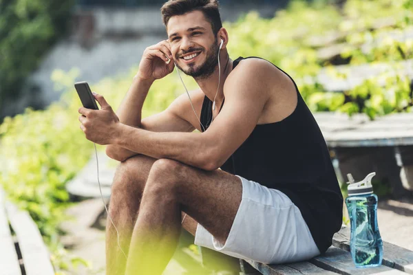 Улыбающийся спортсмен с бутылкой воды слушает музыку со смартфоном и наушниками на скамейке на спортивной площадке — стоковое фото