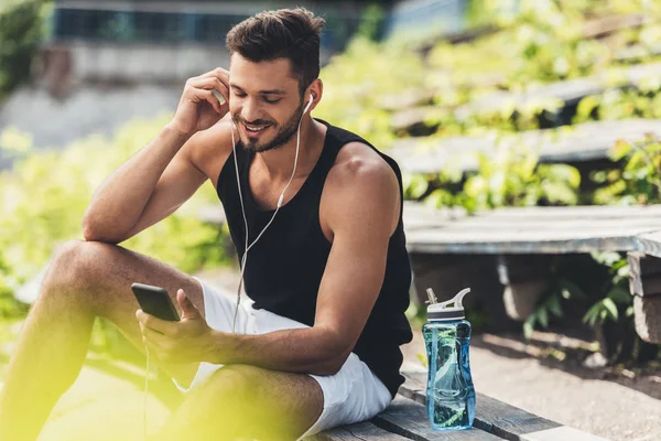 Deportista feliz con botella de agua escuchando música con teléfono inteligente y auriculares en el banco en el parque deportivo - foto de stock