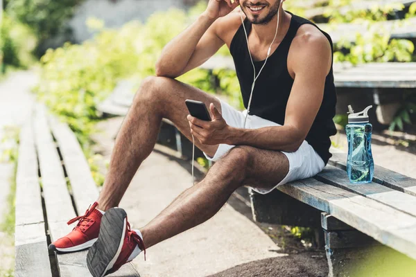 Cropped shot of sportsman with bottle of water listening music with smartphone and earphones on bench at sport playground — Stock Photo
