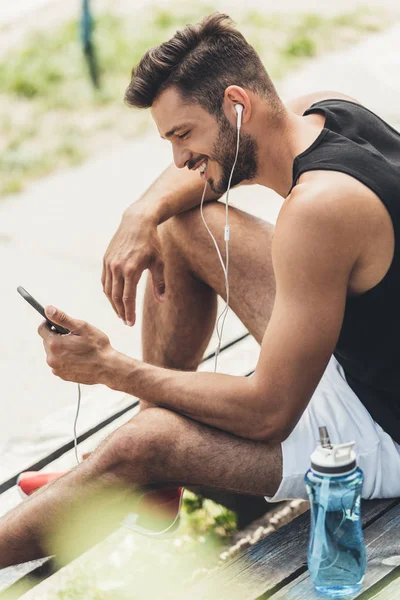 Sportler mit Wasserflasche hört Musik mit Smartphone und Kopfhörer auf Bank auf Sportplatz — Stockfoto