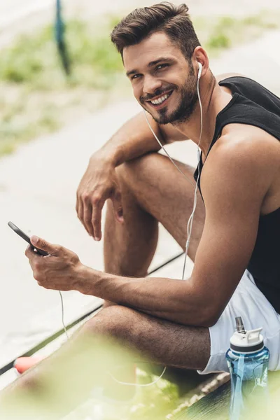 Junger lächelnder Mann mit Wasserflasche hört Musik mit Smartphone und Kopfhörer auf Bank — Stockfoto