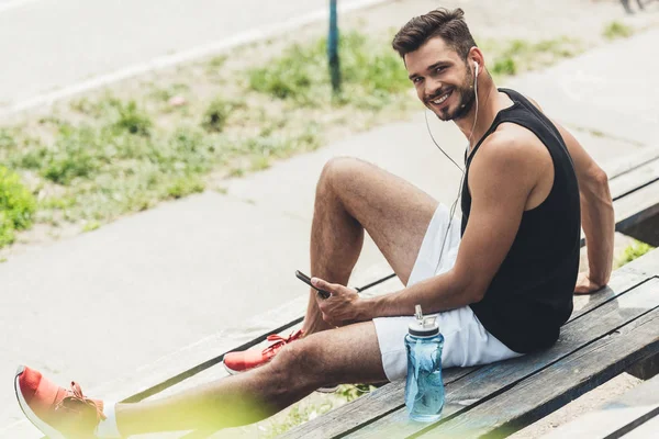 Молодой улыбающийся человек с бутылкой воды слушает музыку со смартфоном и наушниками на скамейке — стоковое фото