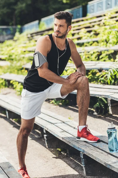 Joven deportista en auriculares con teléfono inteligente en caso de correr brazalete haciendo ejercicio en el banco en el patio de recreo deportivo - foto de stock