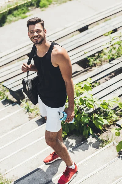 Hochwinkelaufnahme eines glücklichen Sportlers, der eine Flasche Wasser und eine Tasche für Sportgeräte auf der Treppe am Sportplatz in der Hand hält — Stockfoto
