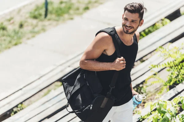 Sportivo sorridente che tiene una bottiglia d'acqua e una borsa per attrezzature sportive sulle scale del parco giochi sportivo — Foto stock