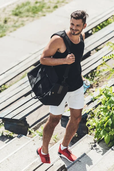 Усміхнений спортсмен, що йде нагору з пляшкою води та сумкою для спортивного обладнання на спортивному майданчику — стокове фото