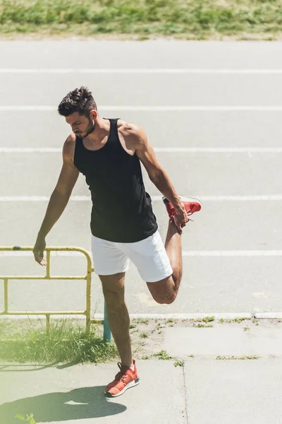 Молодой спортсмен, растянувшийся рядом с беговой дорожкой на спортивной площадке — стоковое фото