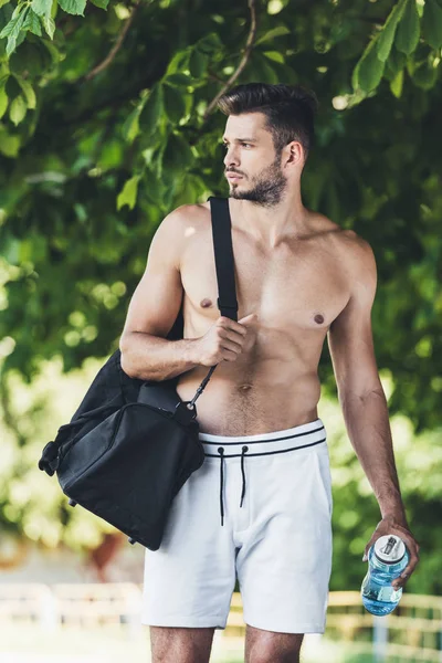 Muscular young man with bag for sport equipment and fitness bottle walking under green tree — Stock Photo