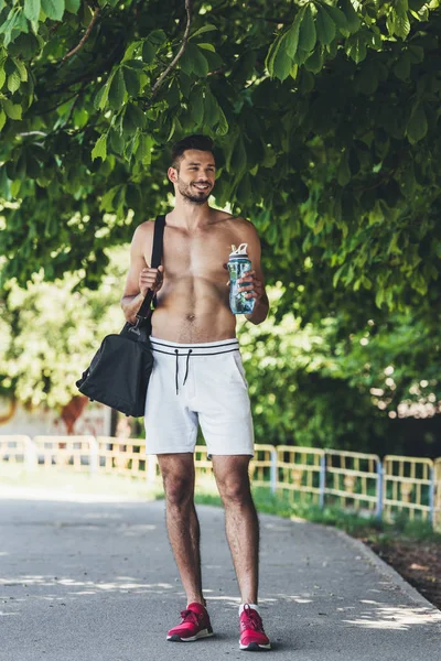 Счастливый молодой человек с сумкой для спортивного инвентаря и бутылкой фитнеса под зеленым деревом — стоковое фото