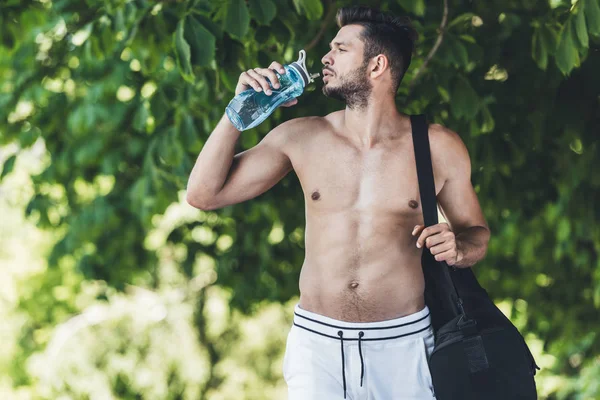 Handsome young man with bag for sport equipment drinking water from fitness bottle — Stock Photo
