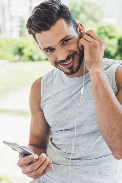 Portrait en gros plan d'un jeune homme heureux écoutant de la musique avec smartphone et écouteurs et regardant la caméra à l'extérieur — Photo de stock
