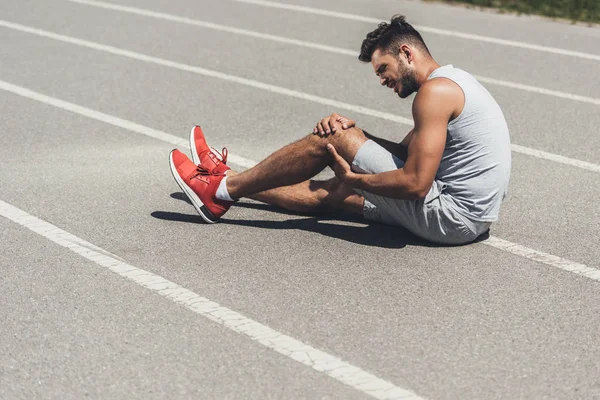 Corredor joven con lesión en la pierna sentado en el suelo de la pista de atletismo - foto de stock