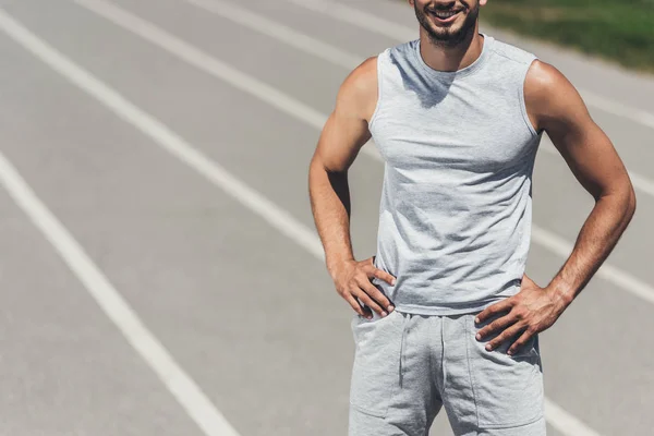 Recortado disparo de sonriente hombre deportivo de pie en pista de atletismo con los brazos akimbo - foto de stock